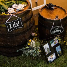 Decor rustic la o nunta outdoor, cu tăblițe "I do" și pozele mirilor așezate pe butoaie de vin.