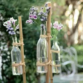 Idee de decor cu flori de camp la o nuntă în aer liber cu sticle simple folosite ca vase de flori legate in vârful unor bete de bambus înfipte în iarbă.