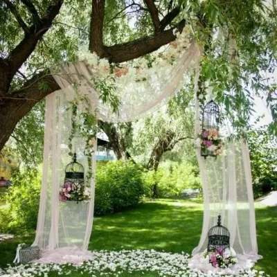 Decor pentru o ceremonie de nuntă în aer liber, cu perdele albe elegante atașate de un copac bătrân, felinare suspendate și petale de flori pe iarbă.
